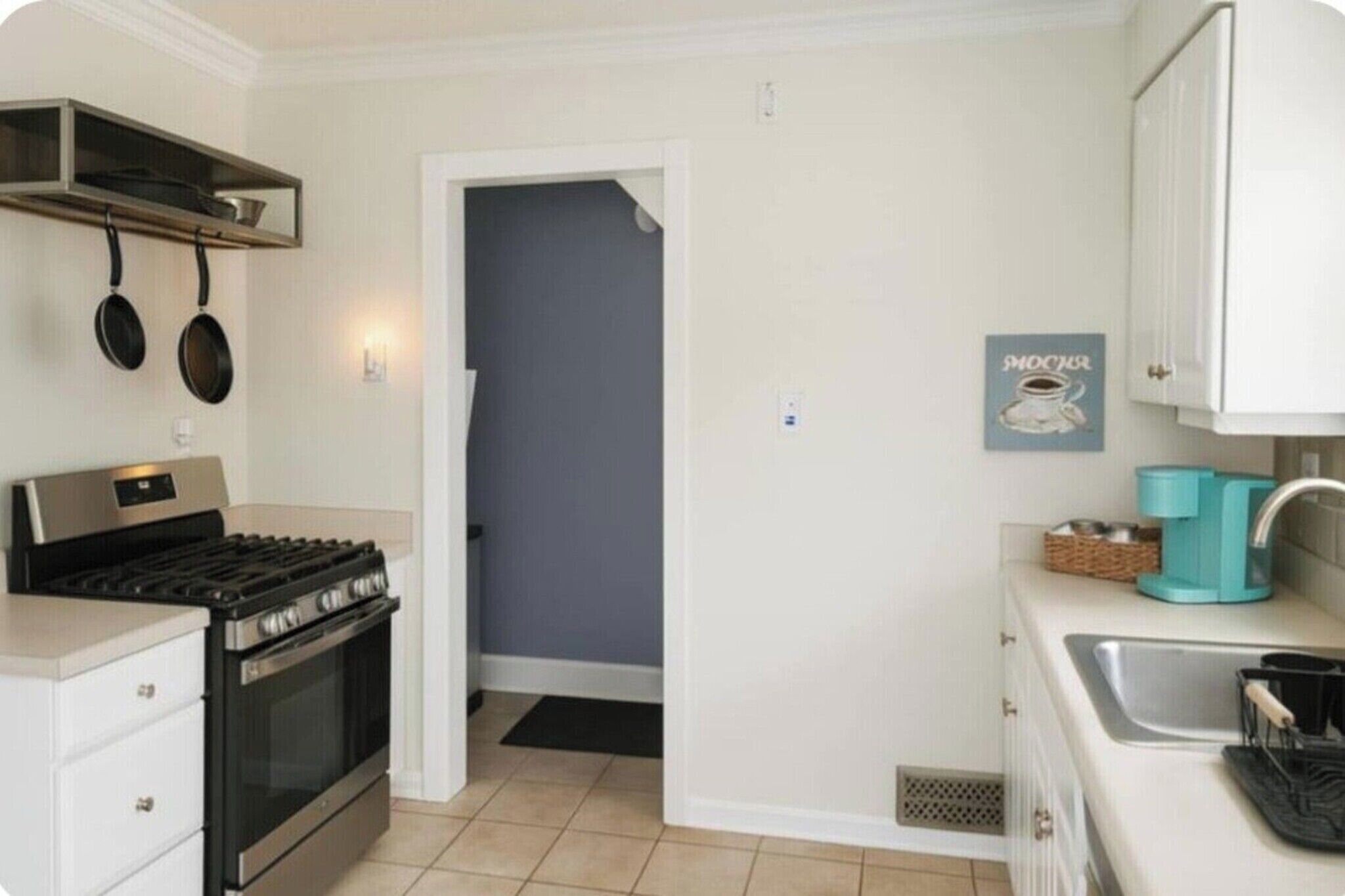 A compact kitchen with a stove, oven, and hanging pots. There's a door leading to another room, a sink with a faucet, and a coffee machine.