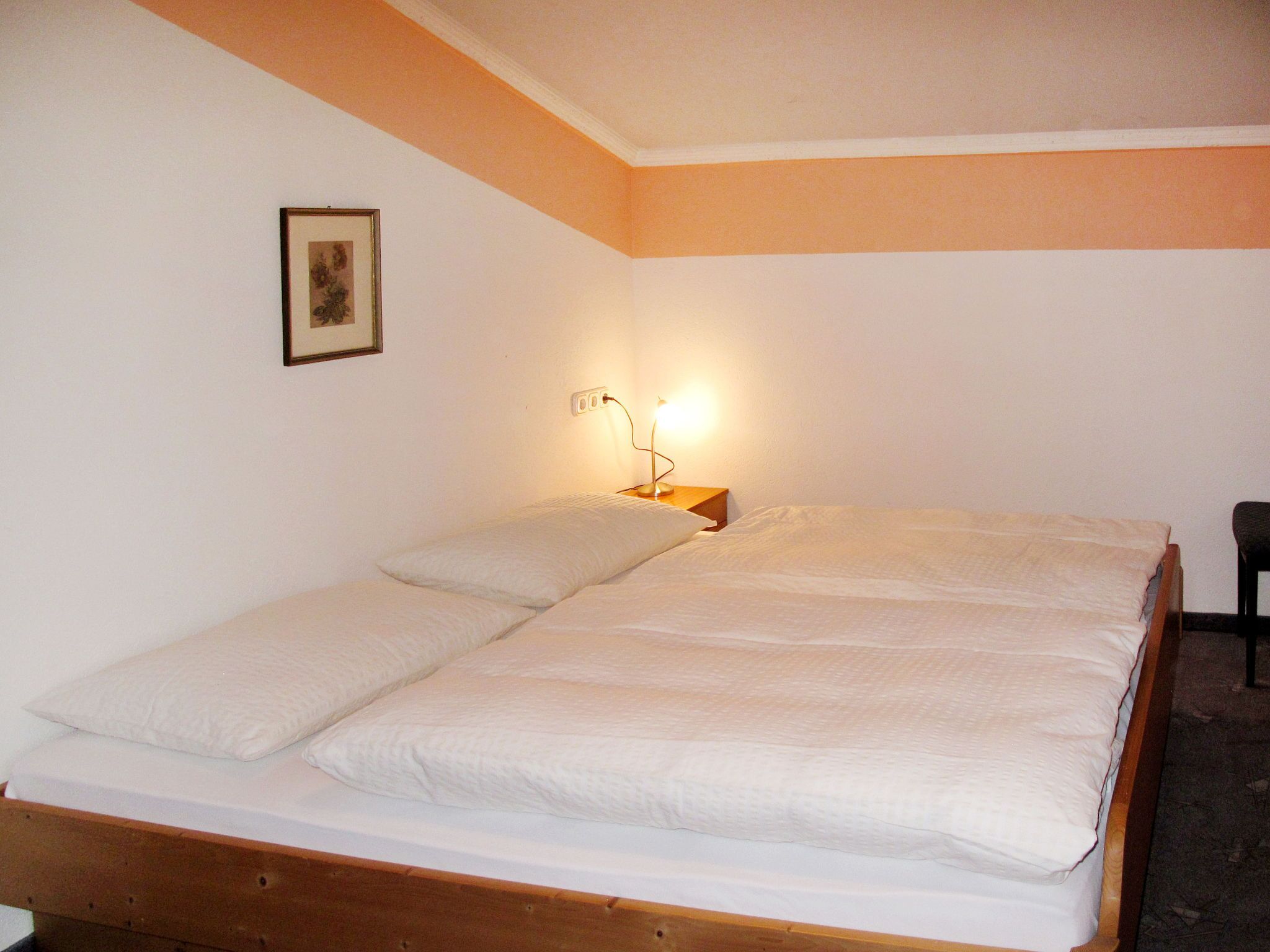 Una cama individual con estructura de madera y ropa de cama blanca en una habitación con paredes blancas y techo inclinado.