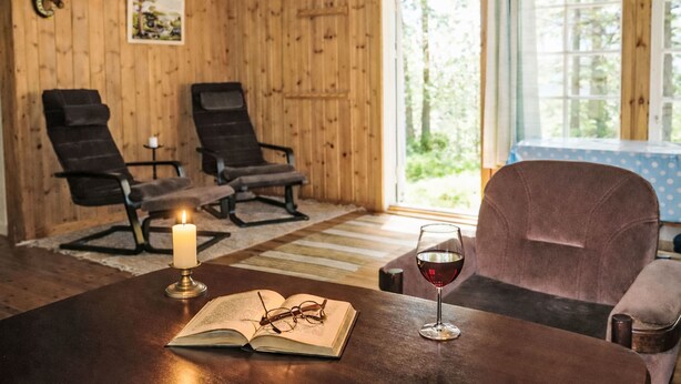 Une pièce chaleureuse aux murs en bois, avec deux chaises noires, un canapé violet, une table sur laquelle se trouvent un livre, des lunettes et un verre de vin rouge.