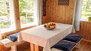 Une table de salle à manger en bois, recouverte d’une nappe blanche, des bancs avec des coussins bleus et un saladier de fruits posé sur la table.