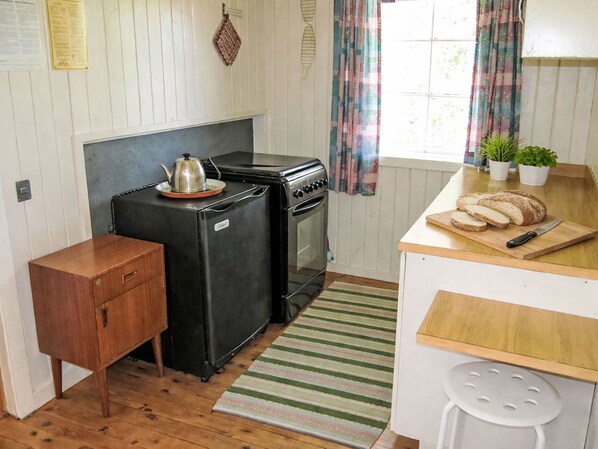 Cocina compacta con refrigerador, estufa y horno negros. Hay un mueble de madera pequeño y una tabla de cortar con pan y un cuchillo en la encimera.