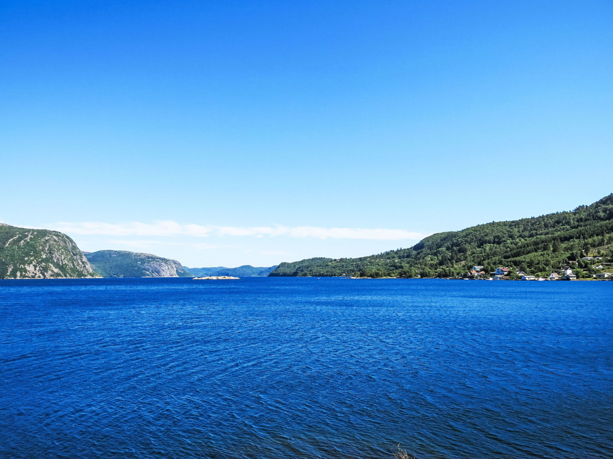 A coastal landscape with a clear blue sea, hills, and a small settlement with houses.