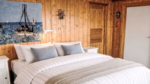 Une chambre à coucher aux murs lambrissés, avec un grand lit, deux tables de chevet, une lampe fixée au mur et un tableau représentant un navire.