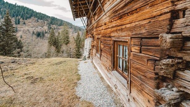 Un chalet rustique en rondins de bois, avec un chemin de gravier menant à l’entrée, entouré d’un paysage montagneux boisé.