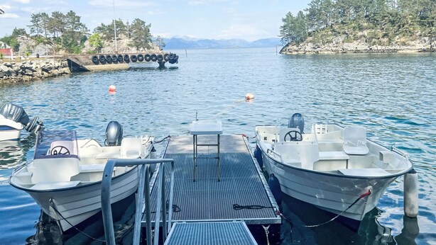 A marina with two boats docked, a metal dock, calm water, and a rocky shoreline with trees. - Gule huset by Interhome (Reksteren)