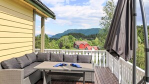 A covered outdoor seating area with a table and cushions, overlooking a scenic landscape with houses and mountains. - Gule huset by Interhome (Reksteren)