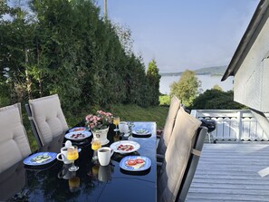 An outdoor dining table with plates of food, cups, and a pitcher, set against a backdrop of greenery and a lake view.