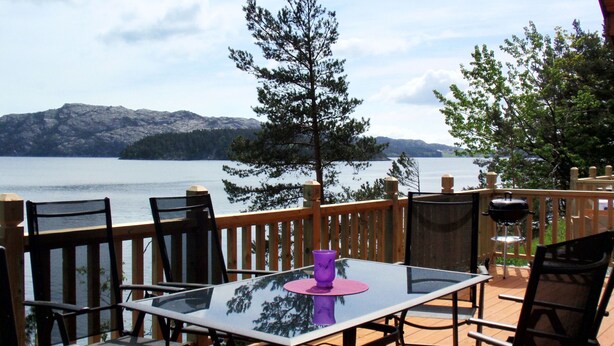Une table à plateau en verre, entourée de chaises, est placée sur une terrasse en bois qui donne sur un lac, avec des montagnes en arrière-plan.