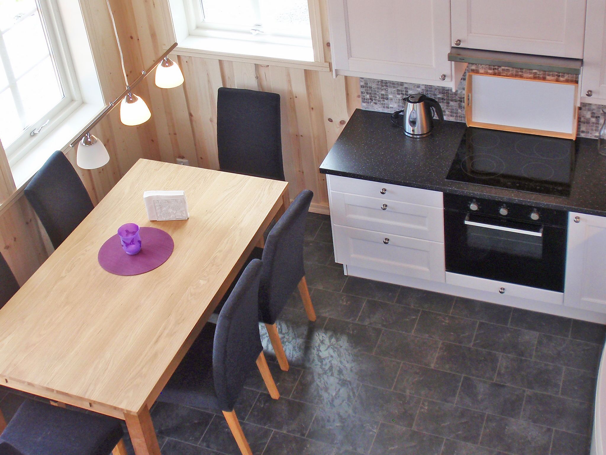 Een kleine keuken met een houten tafel, zwarte stoelen, een waterkoker en een betegelde achterwand.
