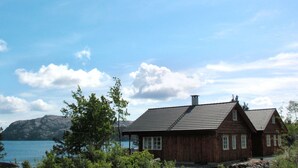 Une cabane en bois avec un toit en pente, située près d’une étendue d’eau, avec des montagnes en arrière-plan.