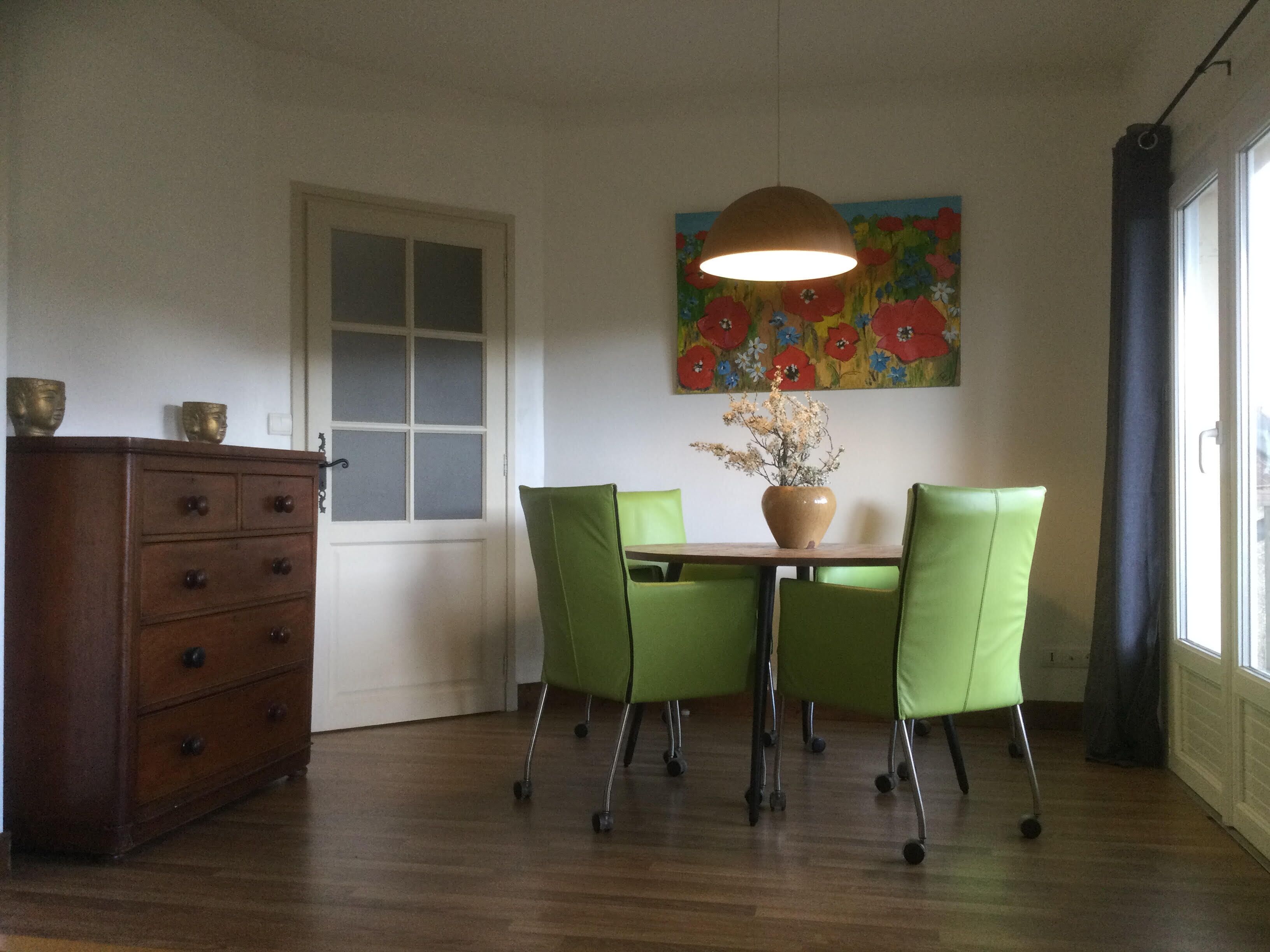 Un coin repas avec une table en bois, quatre chaises vertes, un vase de fleurs, un tableau au mur et un buffet en bois.