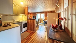 Interior acollidor d'una cabana amb parets i terres de fusta, zona de cuina amb fogó i rentapeus, menjador amb taula i cadires, i nevera amb flors a la part superior.