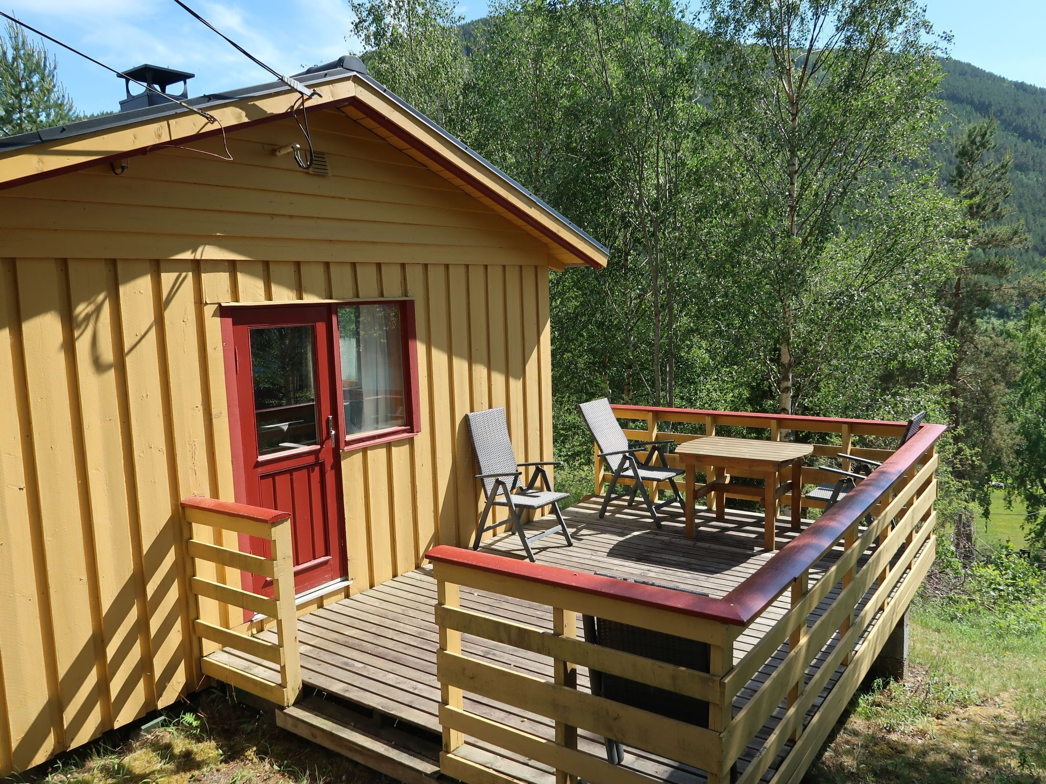 Uma pequena cabana de madeira com porta e janelas vermelhas, um deck com móveis de exterior e vista para uma área arborizada.