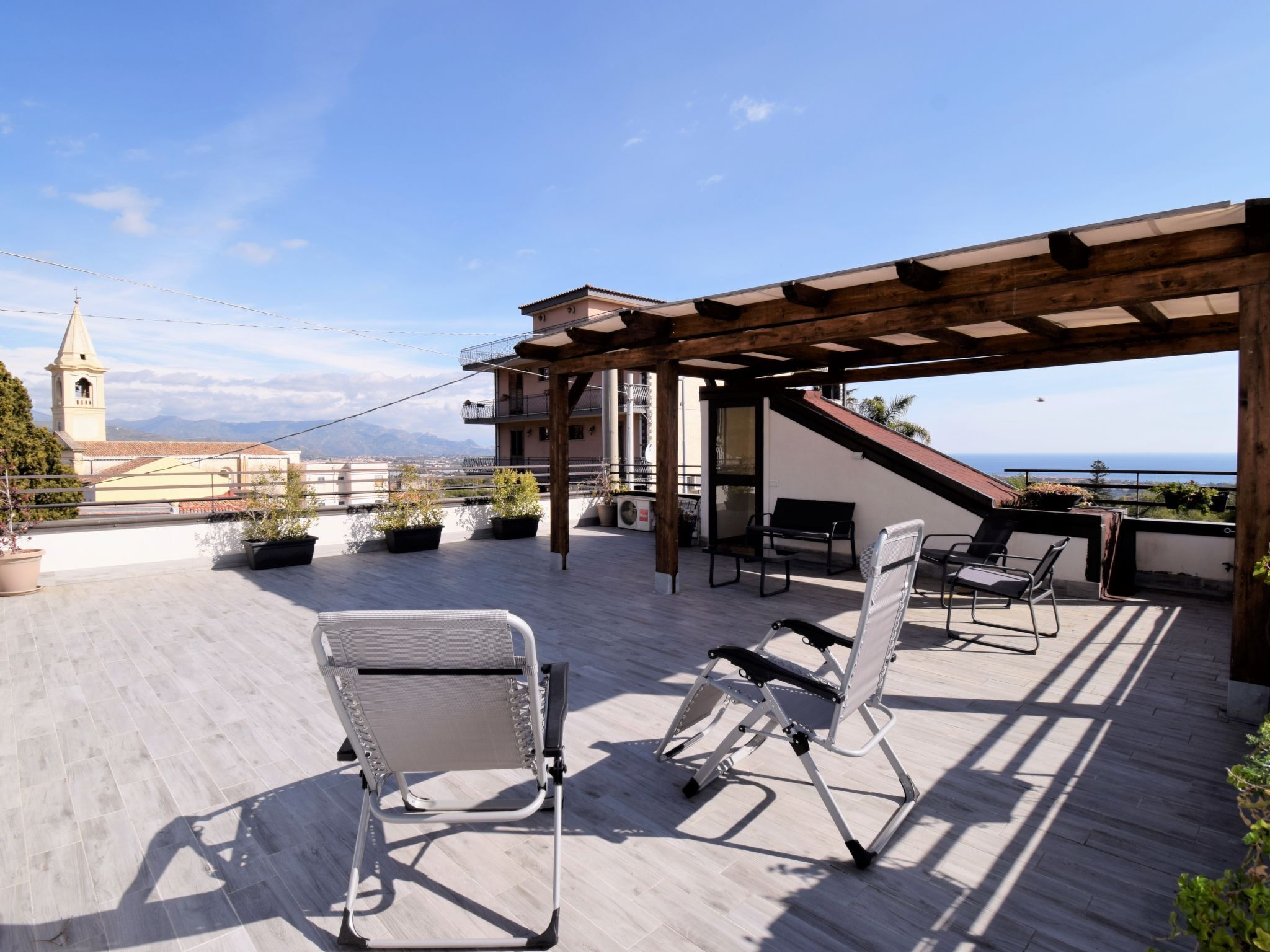 Una terraza en la azotea con mobiliario exterior, una pérgola y vistas al mar y una torre de iglesia.