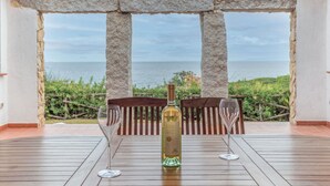 Une terrasse avec une table dressée pour deux personnes, une bouteille de vin et deux verres à vin. La terrasse offre une vue panoramique sur la mer et la verdure environnante.