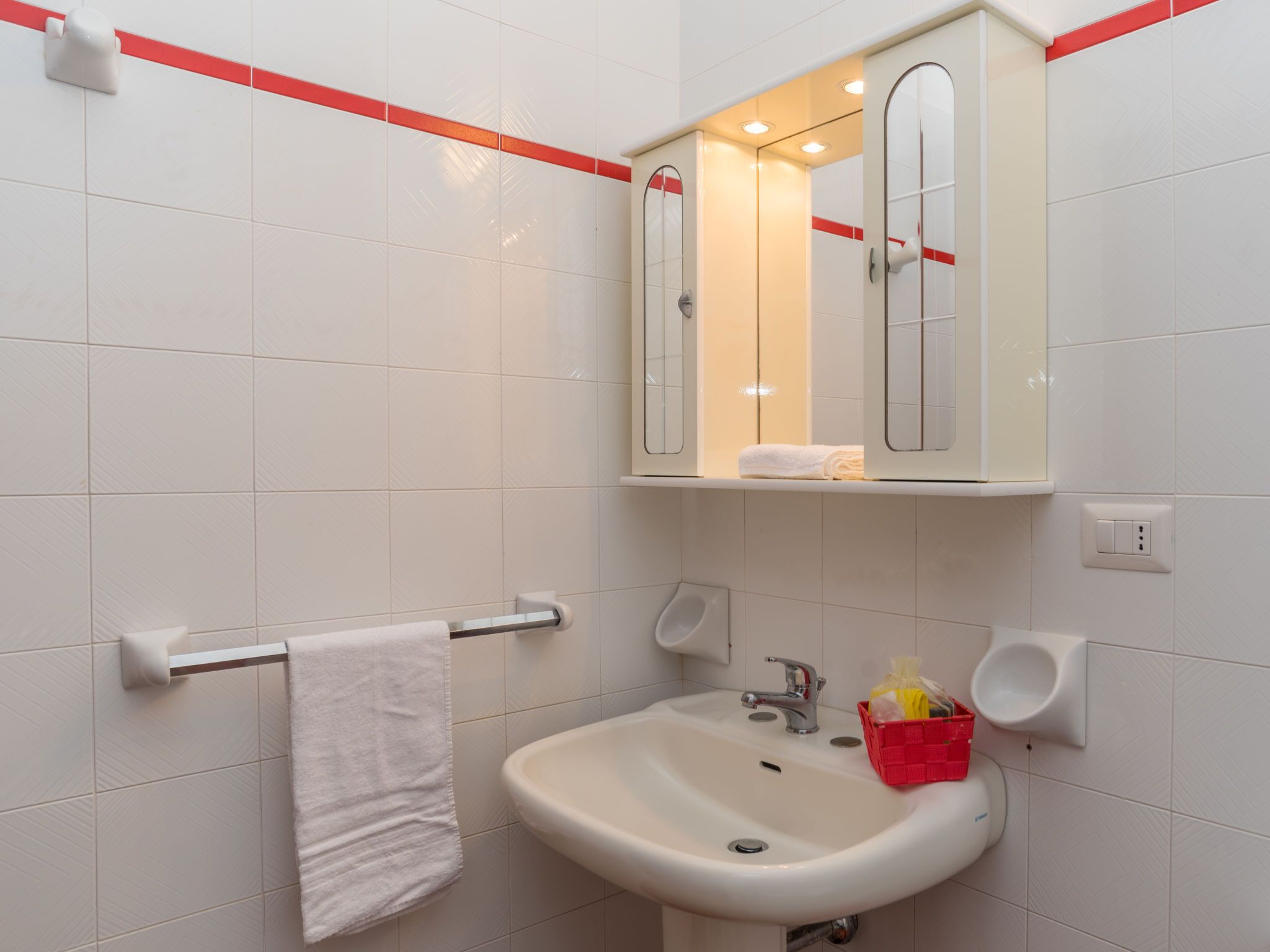 Um banheiro com azulejos brancos, pia, espelho, porta-toalhas e uma cesta vermelha.