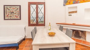 Un coin repas avec une table, des chaises, un saladier de fruits, une bouteille de vin et une vue par la fenêtre.