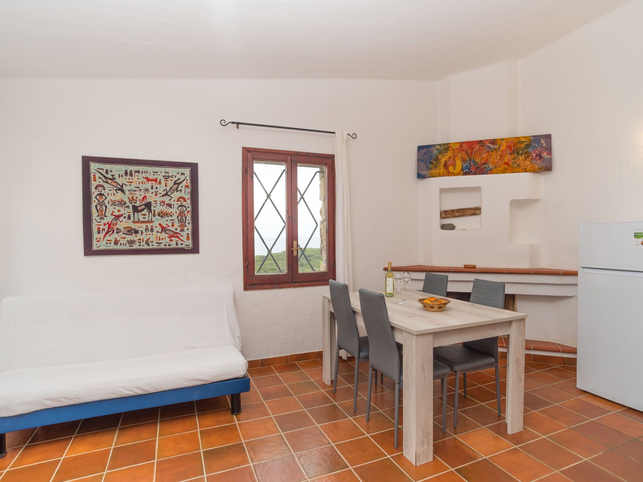 Una habitación compacta con cama, una mesa de comedor con sillas, un ventanal con cortinas, un refrigerador y un cuadro colorido en la pared.