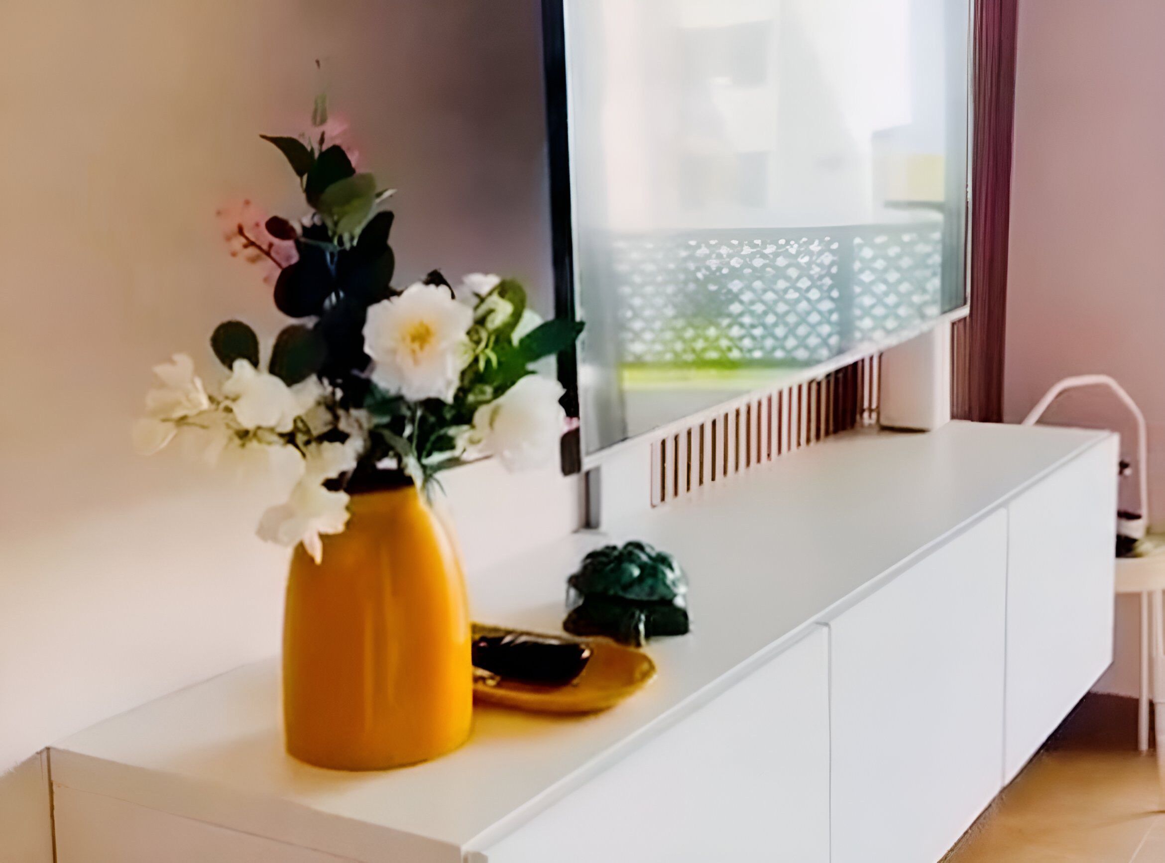 A modern living room with a white console table, a yellow vase with white flowers, a small green plant, and a flat-screen TV mounted on the wall.