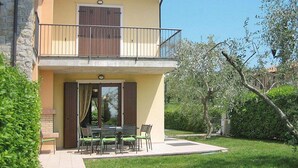 Ein zweistöckiges Gebäude mit einem Balkon, einer Terrasse mit Tisch und Stühlen und einem Garten mit Bäumen und Sträuchern.