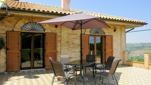 A patio with a table and chairs, a large umbrella, and a brick building with wooden shutters and a chimney.