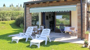 Een overdekt terras met witte plastic stoelen en een tafel, een gebouw van baksteen met een pannendak, en een uitzicht op groen in de verte.