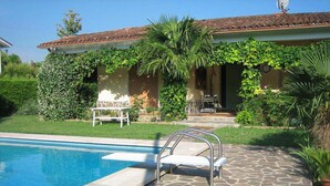 A pool with a stone deck, a white chair, and a covered patio area with a table and chairs. - Vacation home Serena by Interhome (Lazise)