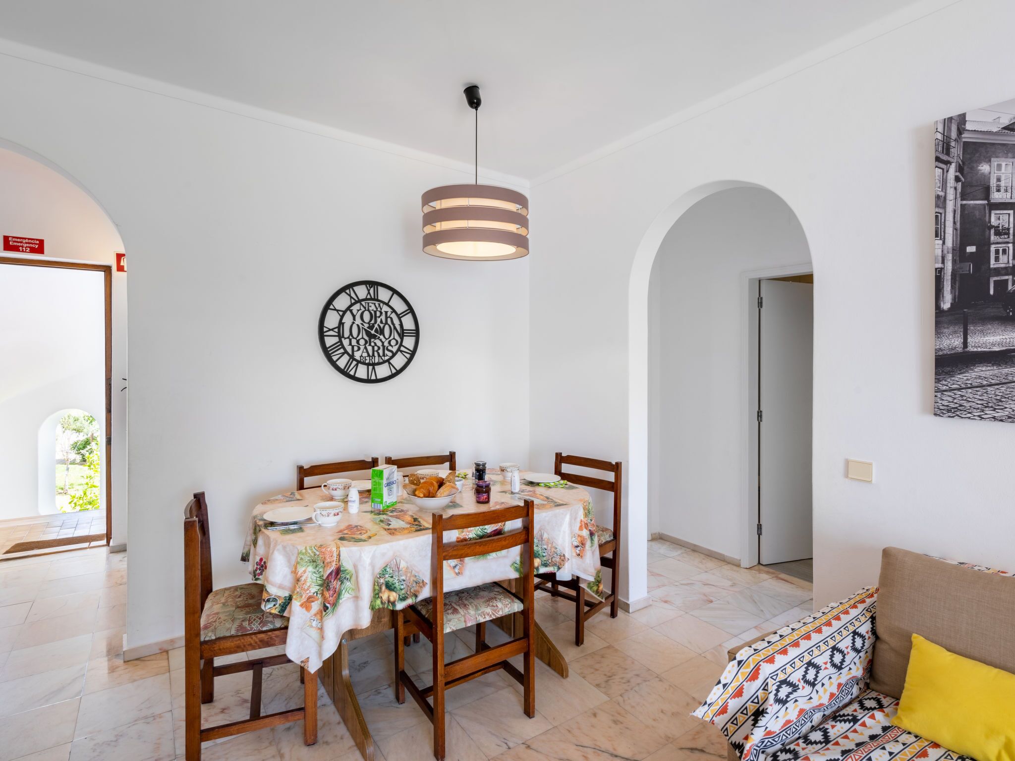 Un comedor con una mesa dispuesta para cuatro, un reloj en la pared y un sofá con un cojín estampado.