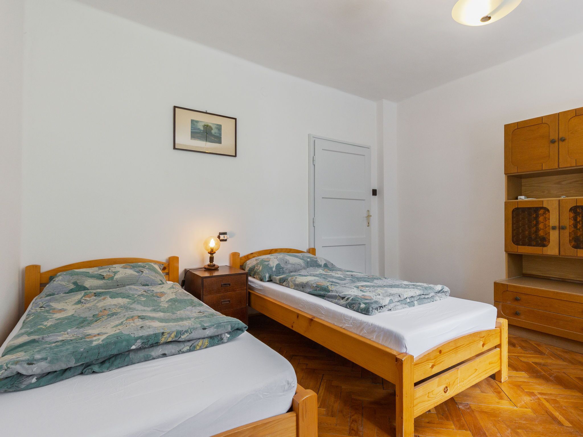 A room with two wooden beds, a small bedside table with a lamp, a white door, and wooden cabinets.
