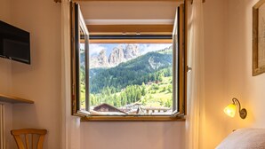 Une chambre avec une grande fenêtre donnant sur les montagnes, un lit recouvert d’une couvre-lit à motifs, une chaise en bois et un téléviseur fixé au mur.