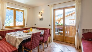 Un coin repas avec une table dressée pour quatre personnes, des chaises à carreaux rouges, un banc et une vue sur une terrasse en bois et les montagnes, le tout visible à travers de grandes fenêtres.