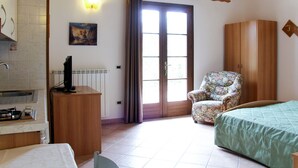 Ein Zimmer mit einem Bett, einem Sessel, einem Kleiderschrank, einem Heizkörper, einem Gemälde und einem Fenster mit Vorhängen.