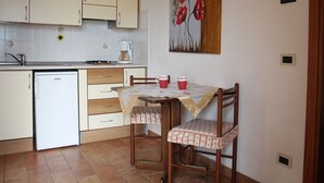 Een compacte keuken met een koelkast, fornuis en koffiezetapparaat. Er staat een eettafel met twee stoelen en er hangt een schilderij aan de muur.
