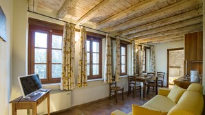 A room with wooden ceiling beams, a dining table with chairs, a sofa, a laptop on a desk, and large windows with curtains.