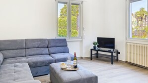 En moderne stue med en grå hjørnesofa, et lille sofabord med en vinflaske og glas, et fjernsyn på et stativ, samt store vinduer med udsigt til træer og bygninger.