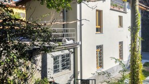 A multi-story building with a balcony, a window with a metal grille, and a tree in the foreground. - Federico by Interhome (Dorio)