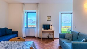 Ein Zimmer mit einem Bett, zwei Sofas, einem Fernseher und einem Schreibtisch. Große Fenster bieten Blick auf einen See und Berge.
