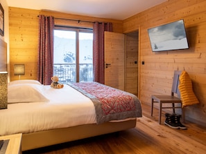 Un dormitorio con una cama, un oso de peluche, un televisor en la pared y una silla con botas.