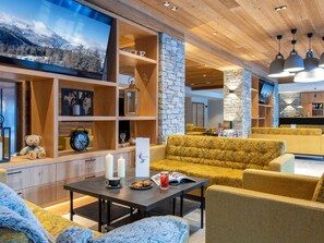 Un vestíbulo de hotel moderno con mobiliario de madera, una pared de acento de piedra y un televisor de pantalla plana grande que muestra una vista nevada de una montaña.