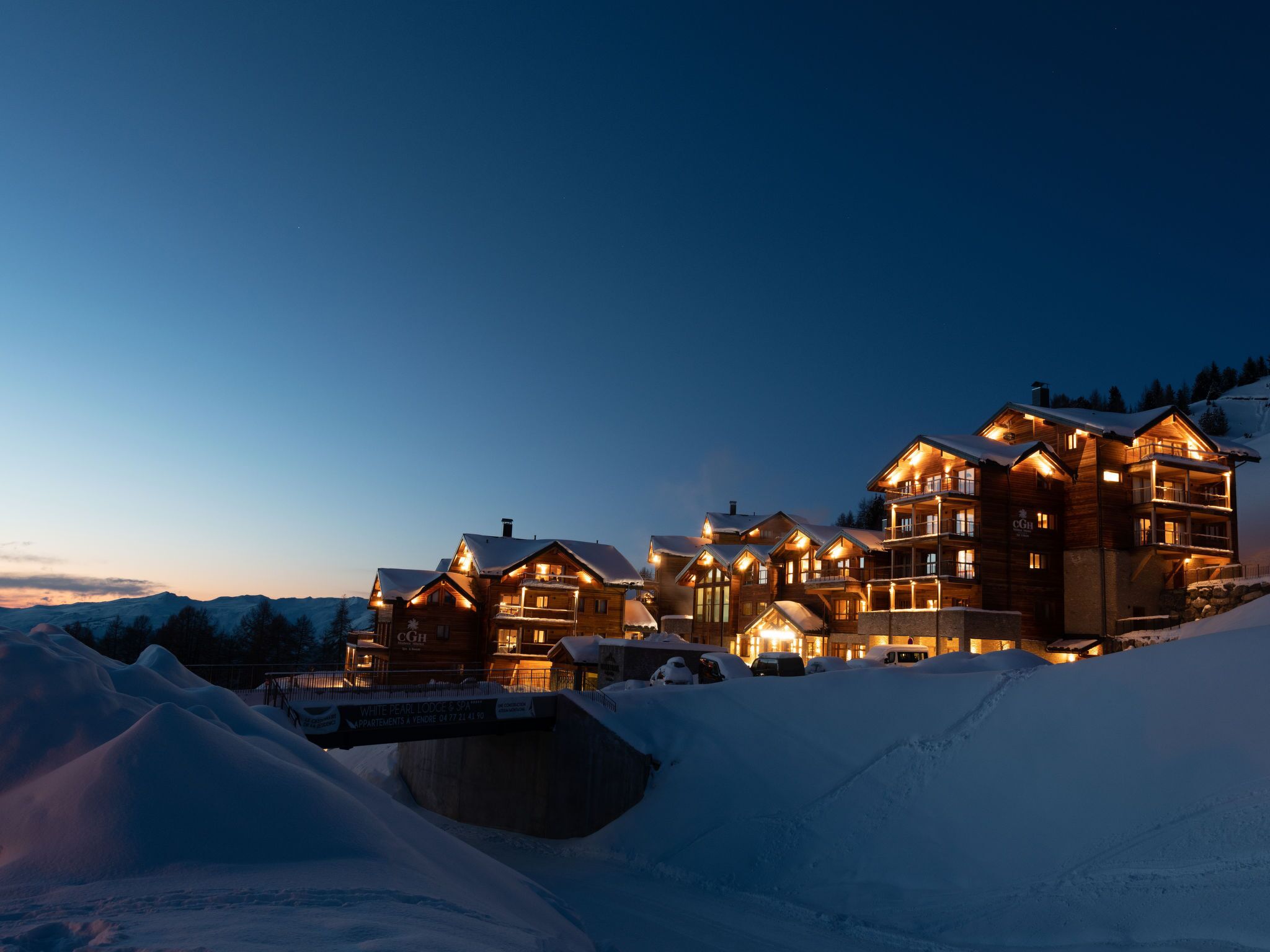 A large, multi-story lodge with a snow-covered roof, illuminated windows, and a sign indicating it is a ski resort.