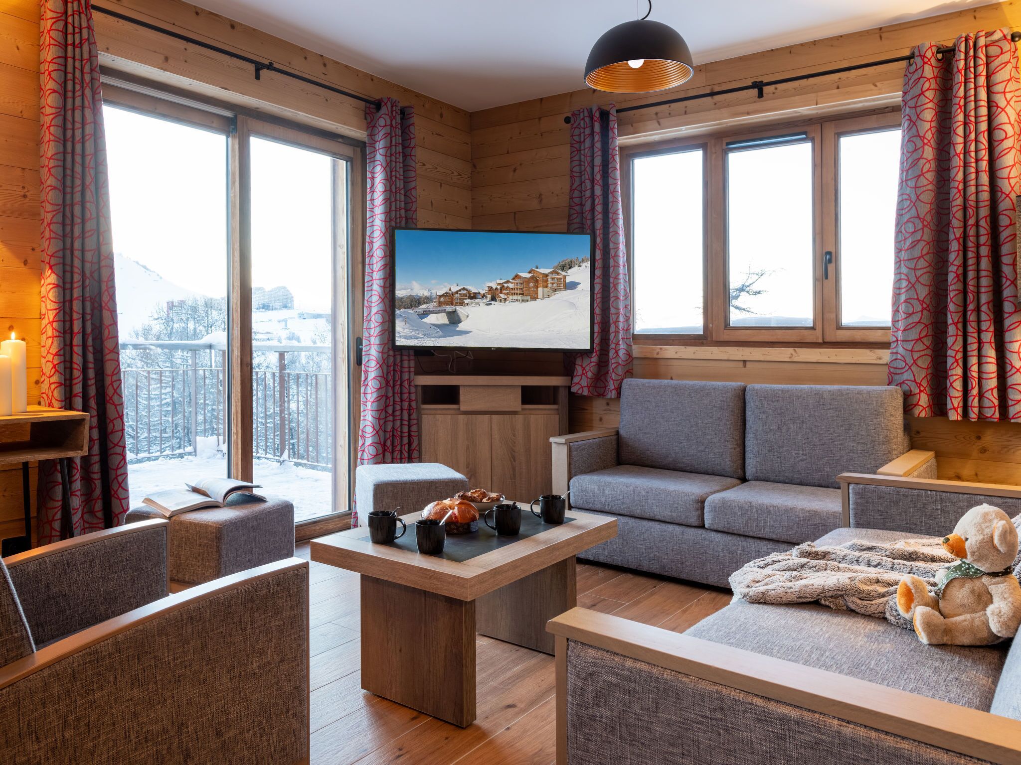 Un salon chaleureux avec un décor intérieur en bois, un téléviseur à écran plat fixé au mur, un canapé, une table basse et un ours en peluche posé sur l’accoudoir.