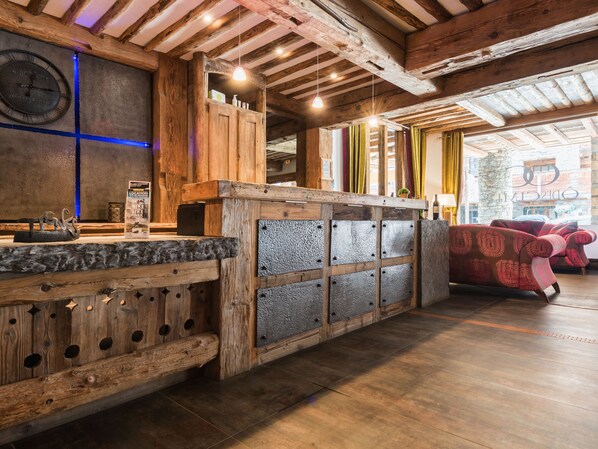 A rustic interior with exposed wooden beams, a stone wall with a clock, a bar area with stools, and a view of the outside through large windows.