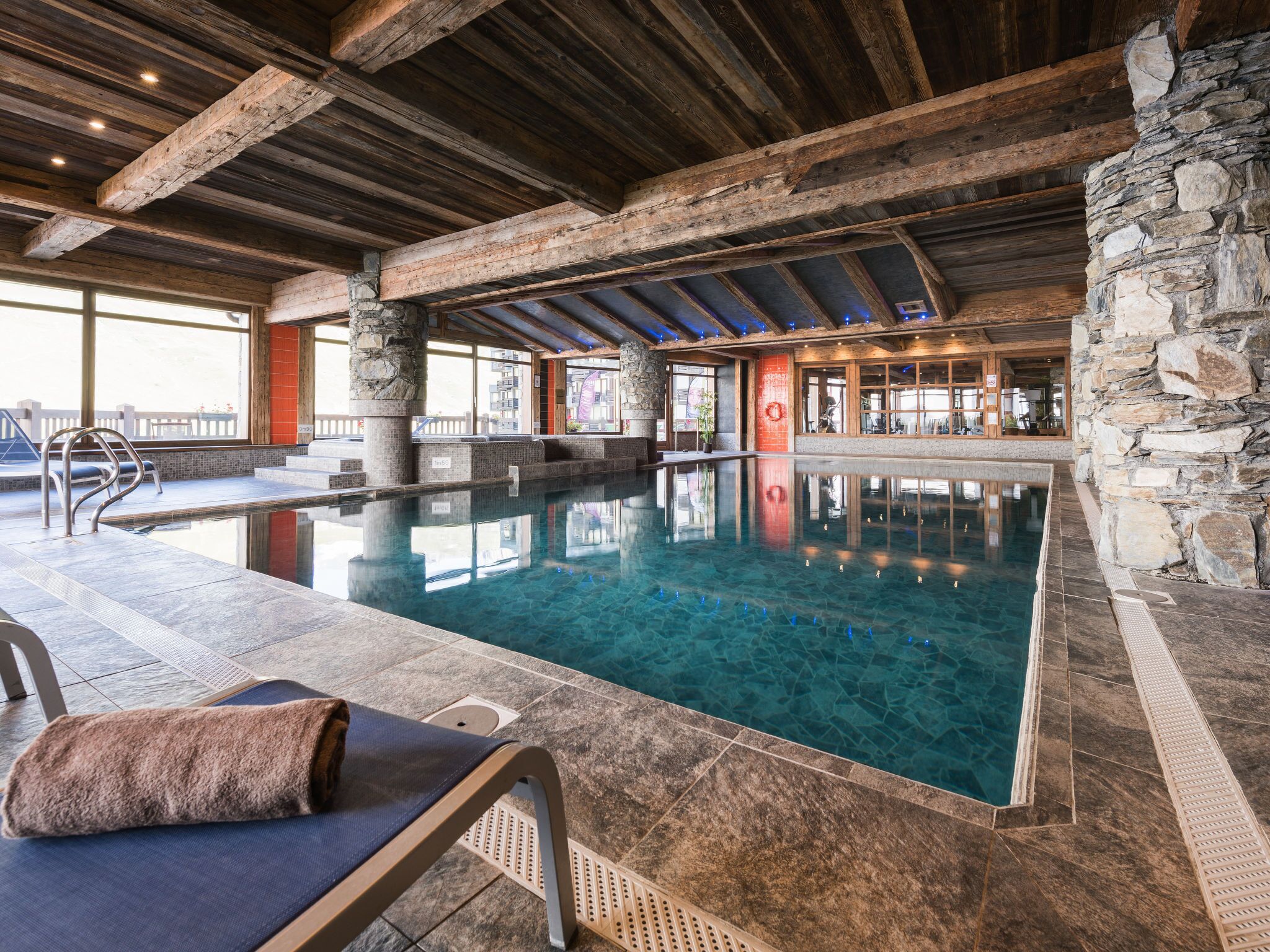 An indoor swimming pool with stone walls and wooden beams, a lounge area with a towel and chair, and a view of the outside.