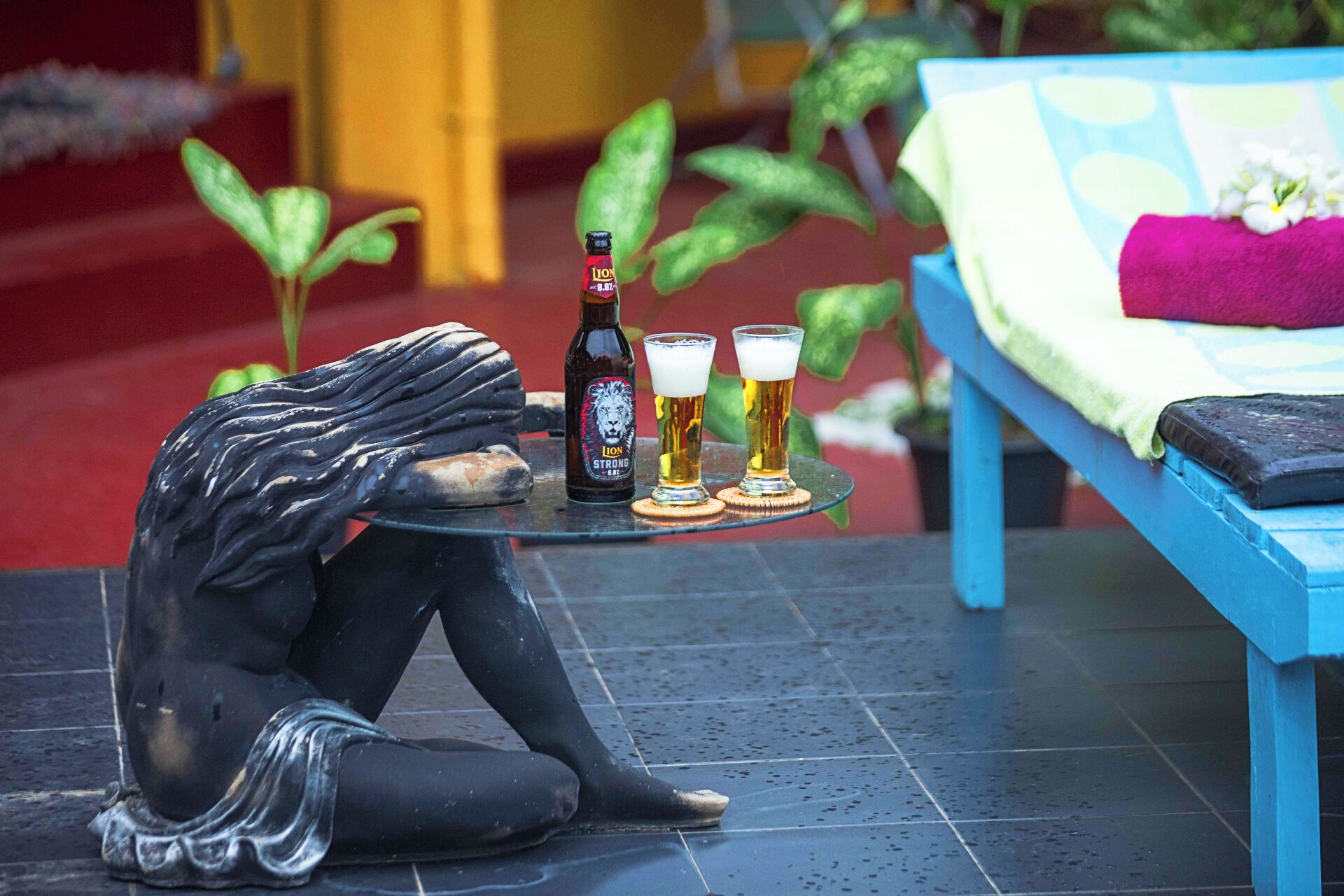 A statue of a person in a dramatic pose on a tiled floor, with a bottle and two glasses of beer on a small table beside it.