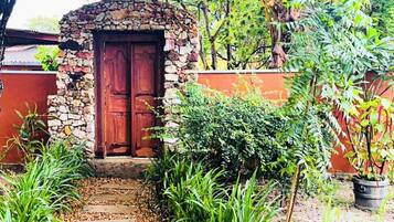Una entrada de piedra con puerta de madera, rodeada de abundante vegetación y un camino de grava.