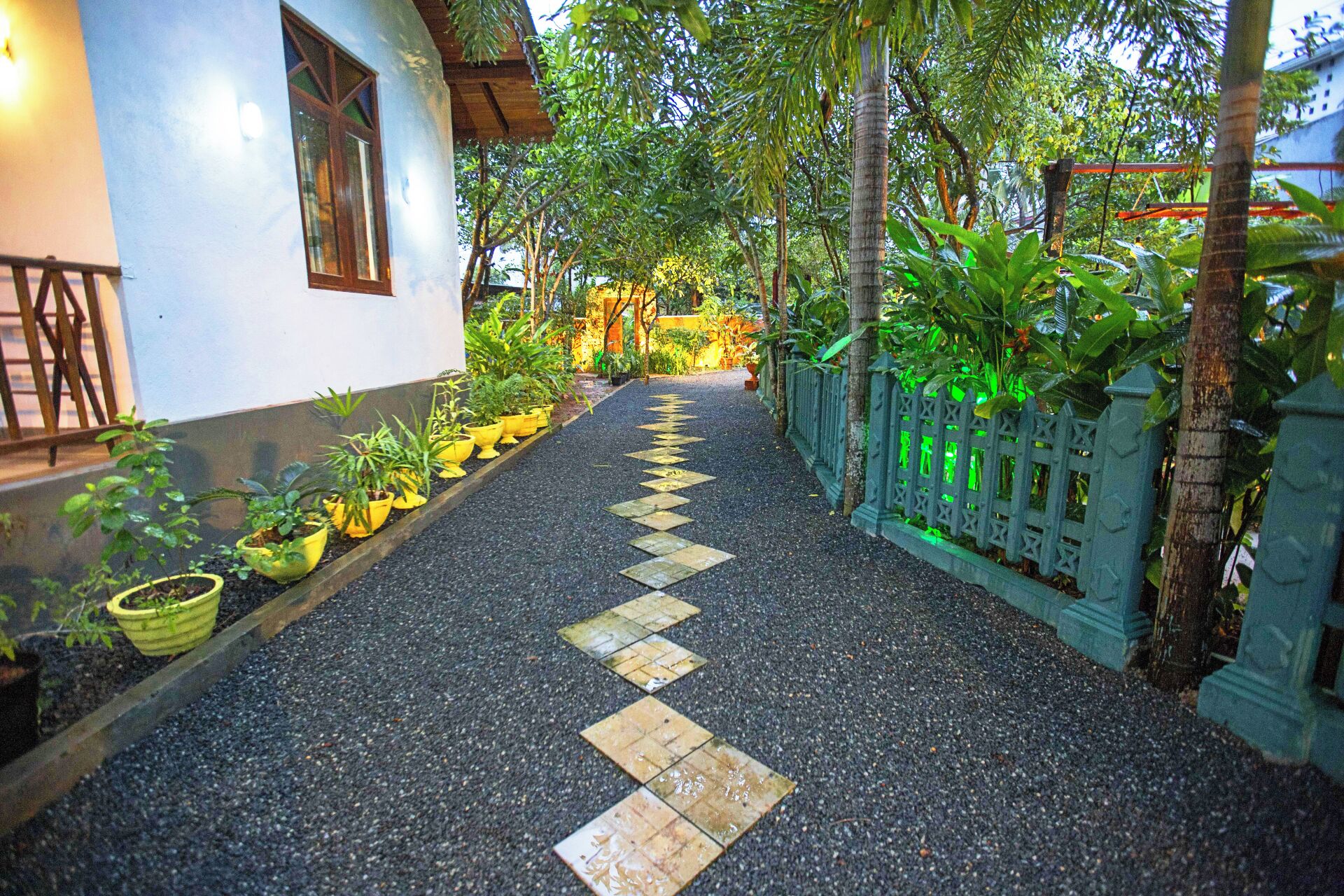 Un chemin bordé de plantes en pot mène à un bâtiment à la façade blanche, doté d’une fenêtre.