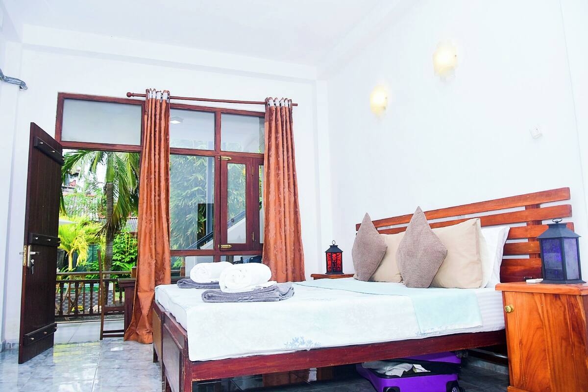 A bedroom with a wooden bed, a nightstand, a lamp, and a view of greenery through a window with curtains.