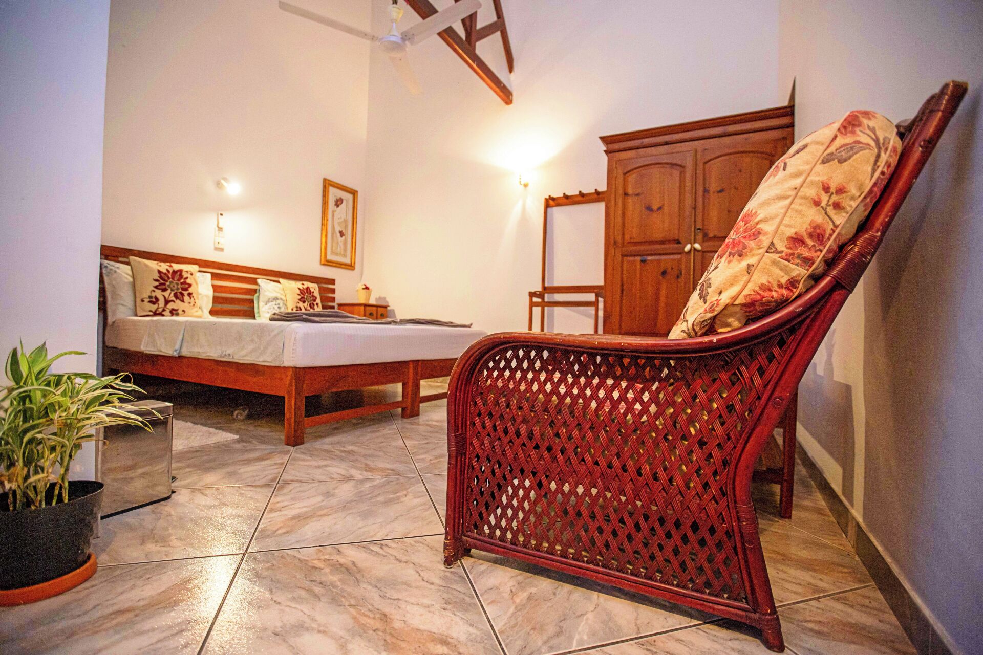 A room with a bed, a wicker chair, a cabinet, and a potted plant.