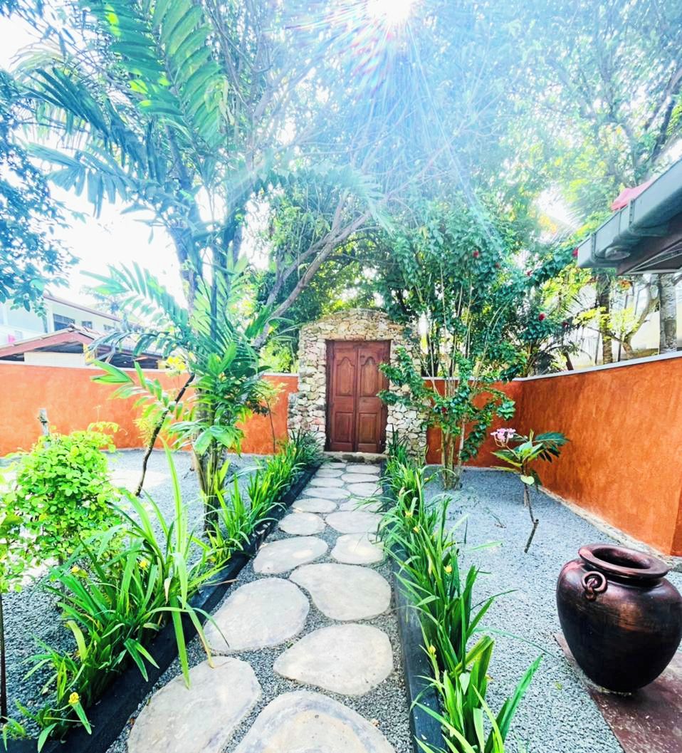 A garden pathway leading to a wooden door, surrounded by lush greenery and a stone wall.