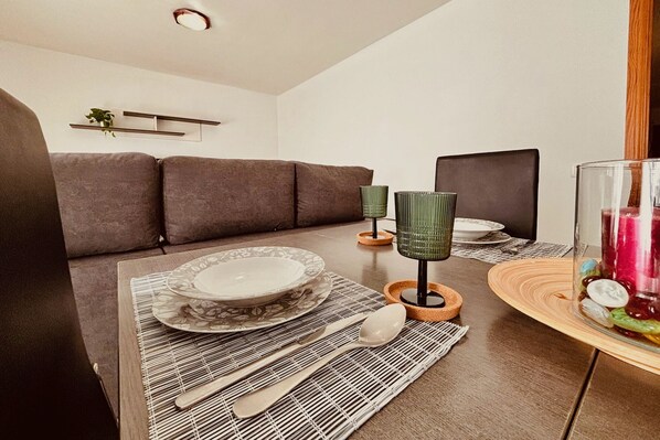 Un coin repas avec une table dressée pour deux personnes, un canapé en arrière-plan et une étagère murale agrémentée d’une plante.
